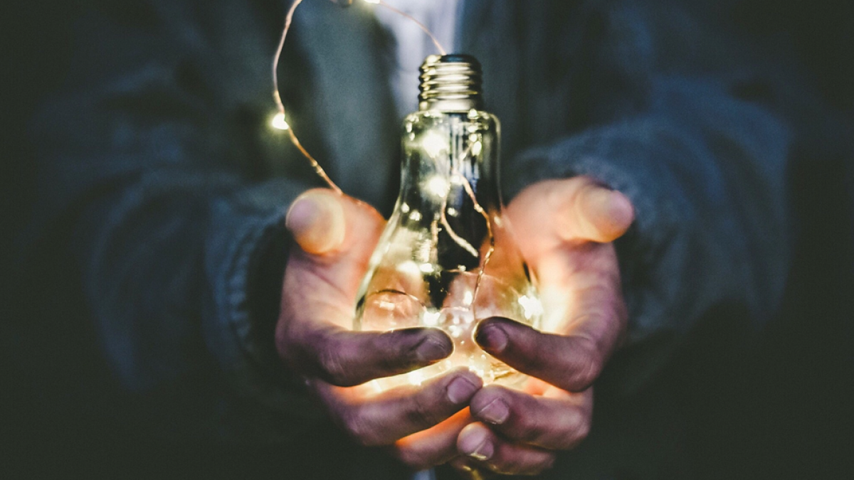 Close-up photograph of a person holding a lightbulb in their cupped hands; the lightbulb is illuminated by a string of smaller lights threaded inside of it.