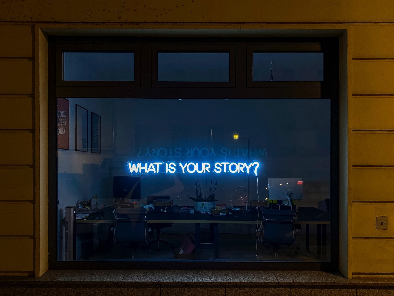 Photograph of a large plate glass window with an illuminated neon sign reading “What is your story” hung behind it.