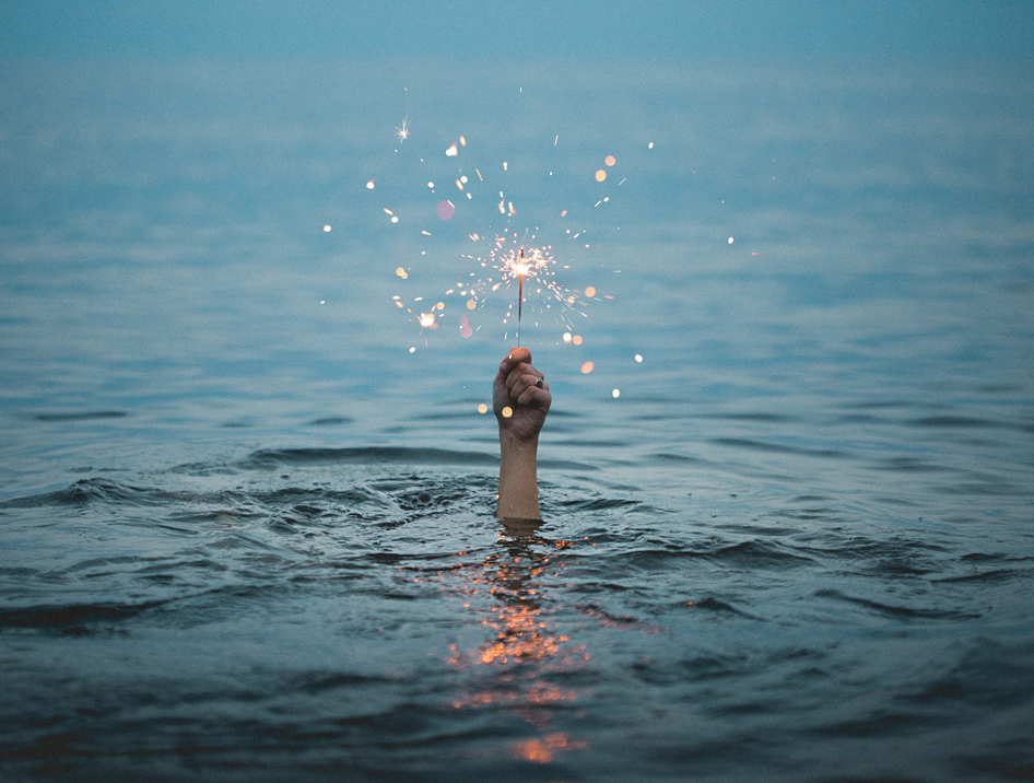 Photograph of hand emerging from beneath the water, holding a lighted sparkler. Image credit: Kristopher Roller/Unsplash