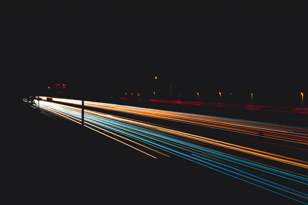 Time-lapse photograph of motor traffic on a highway showing streaks of colored lights from car headlamps and taillights. Image credit: Harrison Kugler/Unsplash