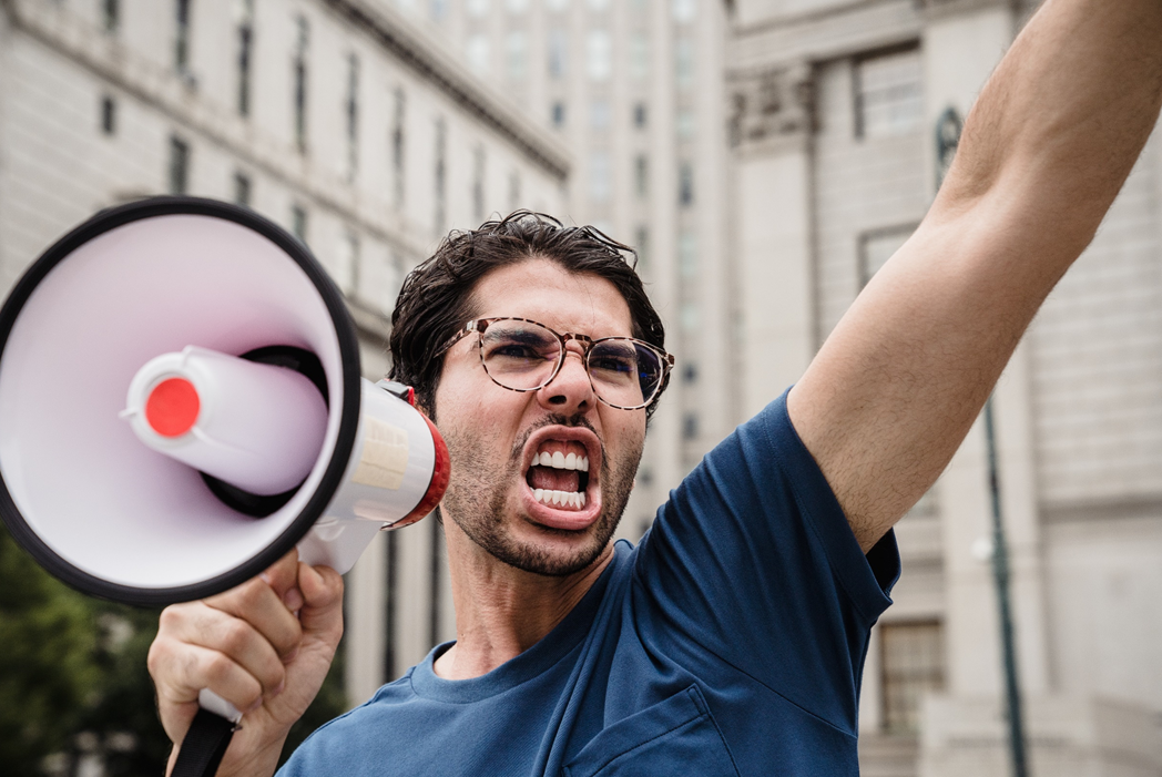 Young man with glasses and angry expression shouting into an electronic bullhorn and raising an arm in the air. Image credit: Lara Jameson/Pexels https://www.pexels.com/photo/city-landscape-man-people-8899224/