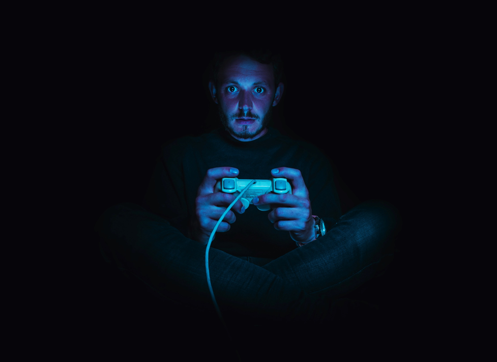 Man sitting in shadow, partly illuminated by blue glow from a monitor or television screen, holding a video game controller, and staring straight at the camera’s perspective. Image credit: Alexander Andrews/Unsplash