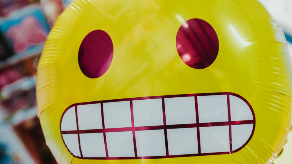 Closeup photo of mylar balloon printed with "grimacing" emoji. Image credit: Bernard Hermant via Unsplash