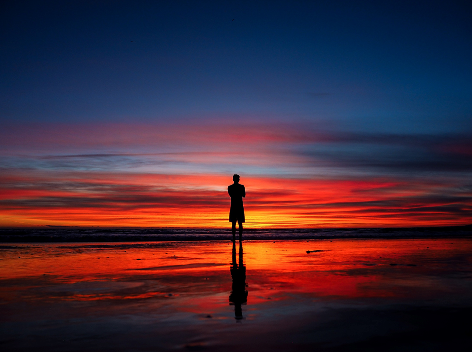 Man standing on a beach with back turned, silhouetted against a colorful sunset sky. Image credit: Maciej Pienczewski/Unsplash