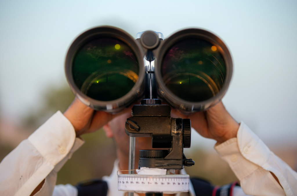 Man facing camera while looking through giant binoculars mounted on a stand or tripod. Image credit: Mostafa Meraji/Unsplash