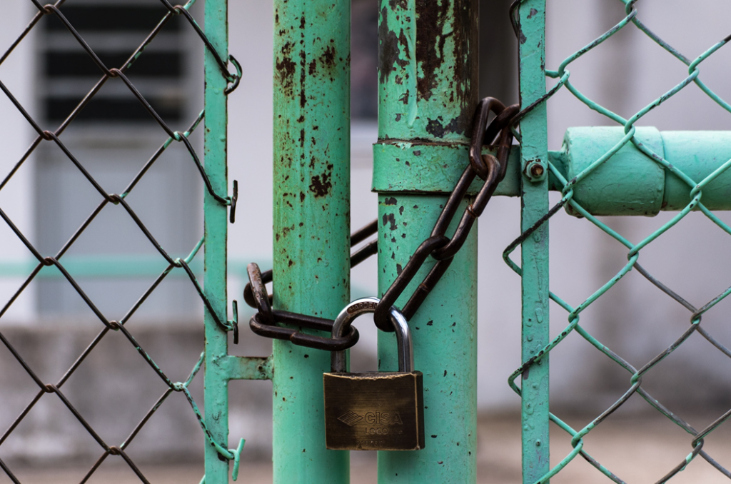 Rusty chain link fence gate closed with chain and padlock, with a building out of focus behind it in the background. Image credit: Jason Blackeye/Unsplash URL: https://unsplash.com/photos/8yYAaguVDgY