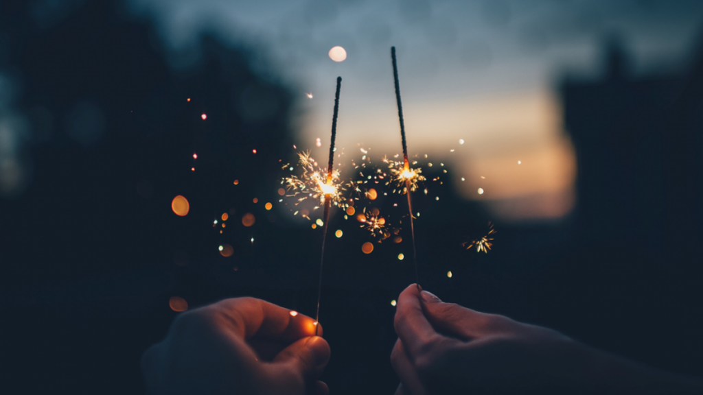 Closeup of two hands holding lit sparklers against a dark background, with sunset visible in the distance. Image credit: Ian Schneider/Unsplash