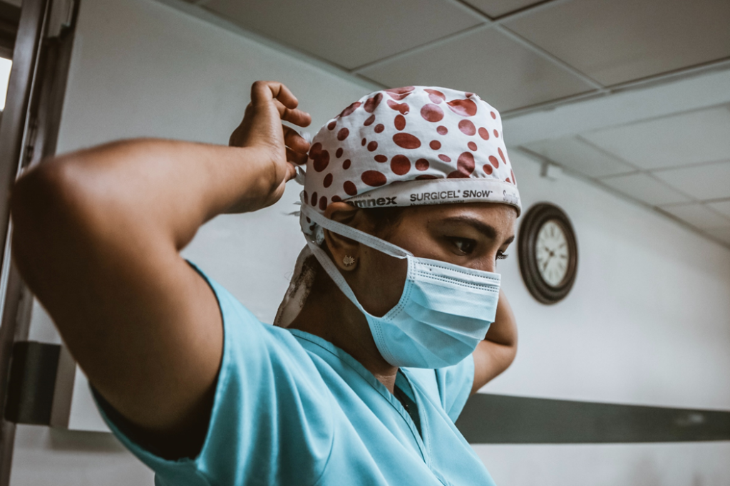 Healthcare worker, dressed in scrubs, with arms raised behind head to adjust surgical mask or head covering. Image credit: SJ Objio/Unsplash