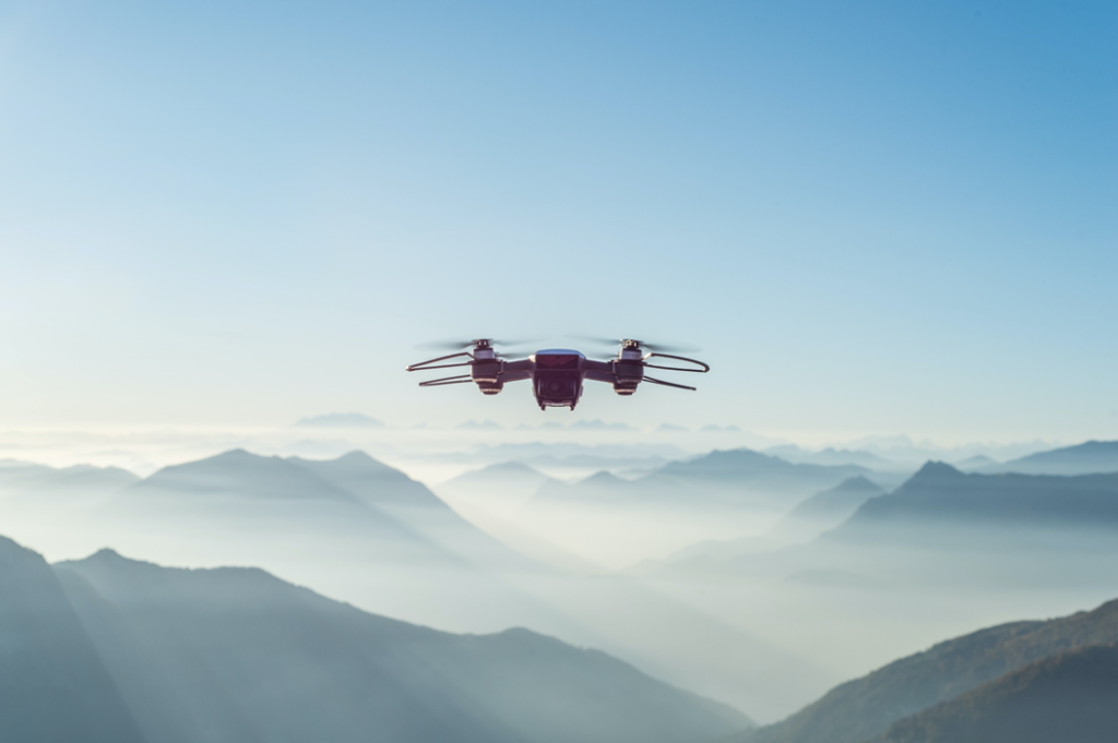 Quadcopter-style drone hovers in midair with mountains, sky, and clouds in the background. Image credit: Alessio Soggetti/Unsplash