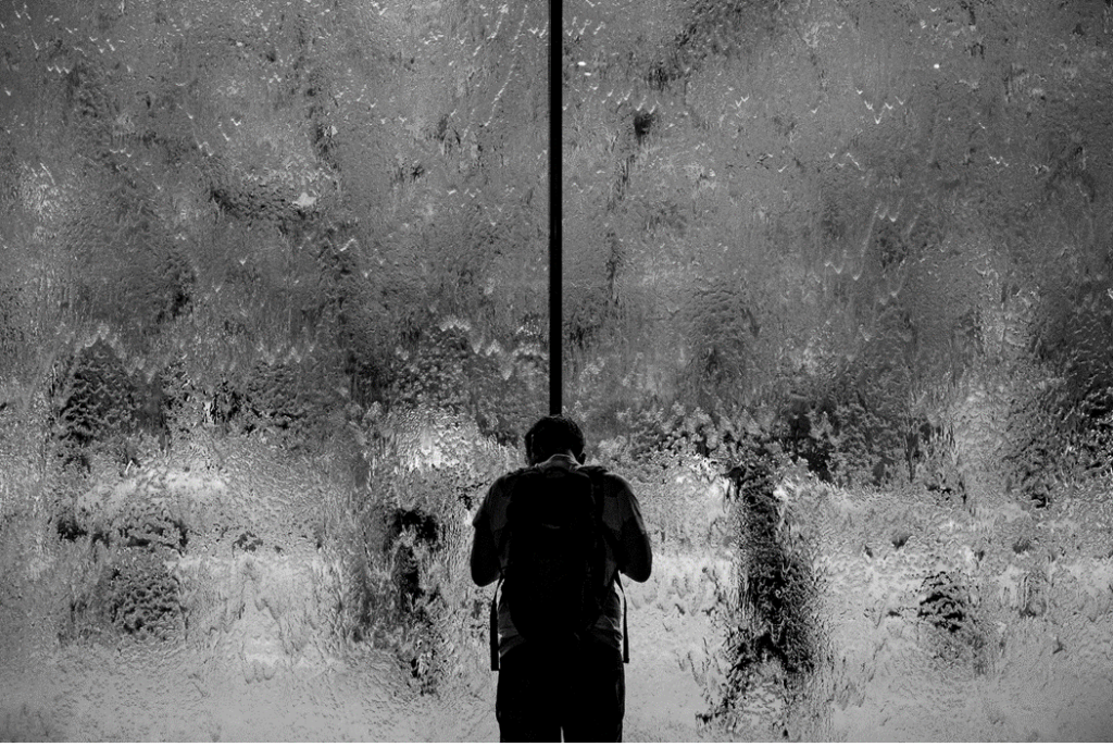 Man in shadow, standing with head bowed and back to camera in front of large glass doors or windows covered with sheets of water as if from a downpour of rain. Image credit: Samuel Austin/Unsplash