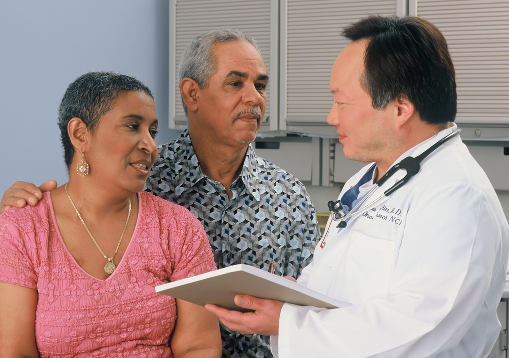 A man and a woman speaking to a doctor in a white lab coat holding a chart, inside a clinic exam room. Image credit: National Cancer Institute