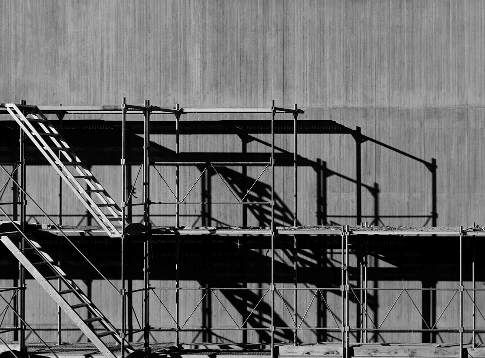 Black and white photograph of metal scaffolding against the side of a building, casting dark shadows on the sunlit wall. Image credit: Ricardo Gomez Angel/Unsplash