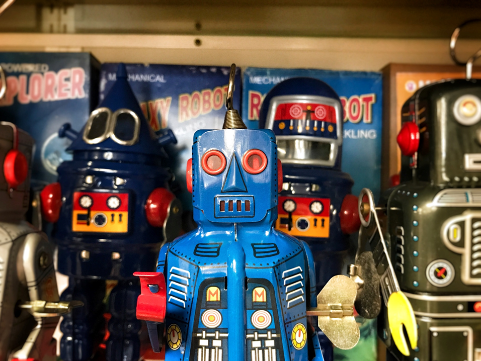 Antique robot toys arranged in a row on display, with one blue robot standing out in front of the rest. Image credit: Craig Sybert/Unsplash
