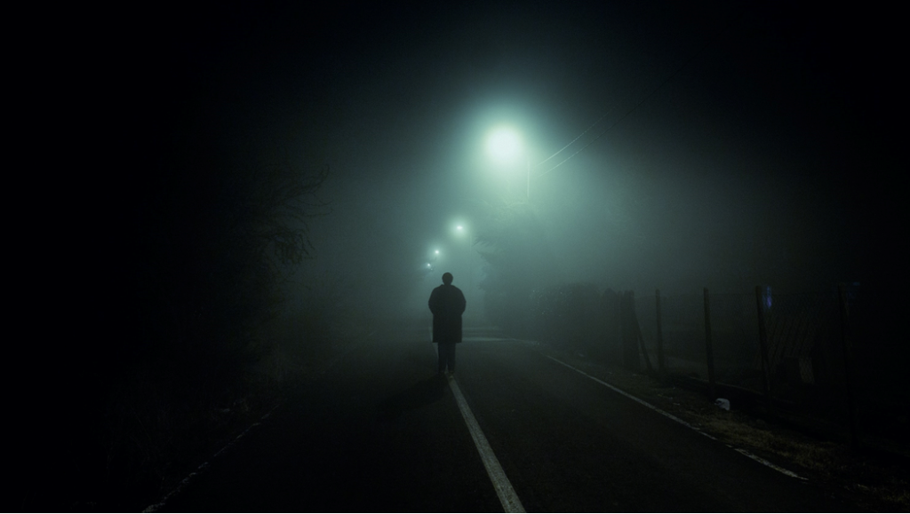 Figure in shadow walking under streetlights on a foggy evening. Image credit: Gabriel/Unsplash