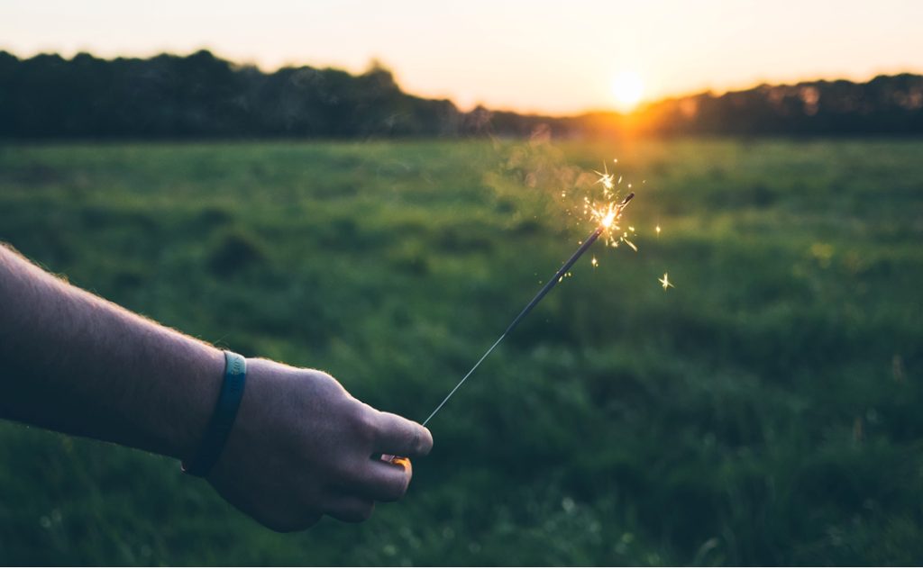 Selective focus photo of a hand holding a lit sparkler in a green field with sun setting in background. Image credit: Jamie Street/Unsplash