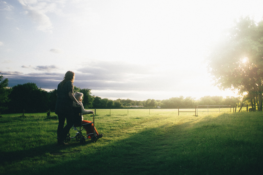 Woman assisting an elderly woman in a wheel chair in a sunlit meadow with a tree in the foreground. Image credit: Domenik Lange/Unsplash