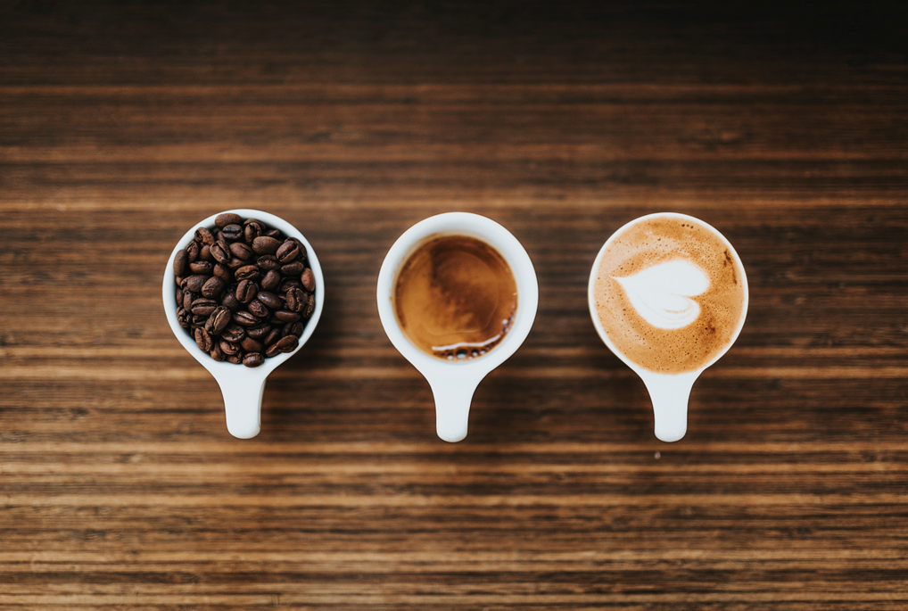 Three white porcelain cups on a wooden surface, photographed from above, containing (from left to right) whole coffee beans, espresso, and a cappuccino with a heart shape drawn in the foam. Image credit: Nathan Dumlao/Unsplash