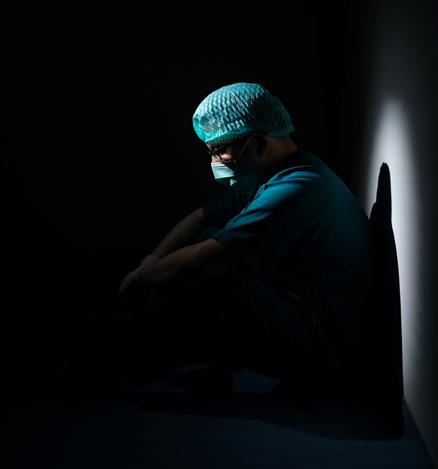 Man dressed in surgical scrubs, standing in profile with eyes cast downward, surrounded by deep shadow. Image credit: Mulyadi/Unsplash