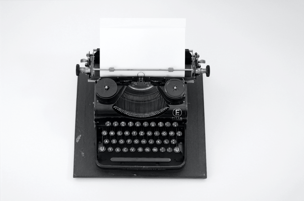 Black mechanical typewriter, viewed from above, with a sheet of blank white paper rolled into the carriage. Image credit: Katrin Hauf/Unsplash