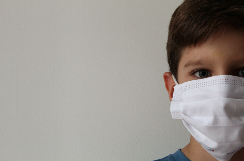 A boy with brown hair and eyes looking directly toward the camera’s perspective, wearing a white surgical mask. Image credit: Lucia Macedo/Unsplash