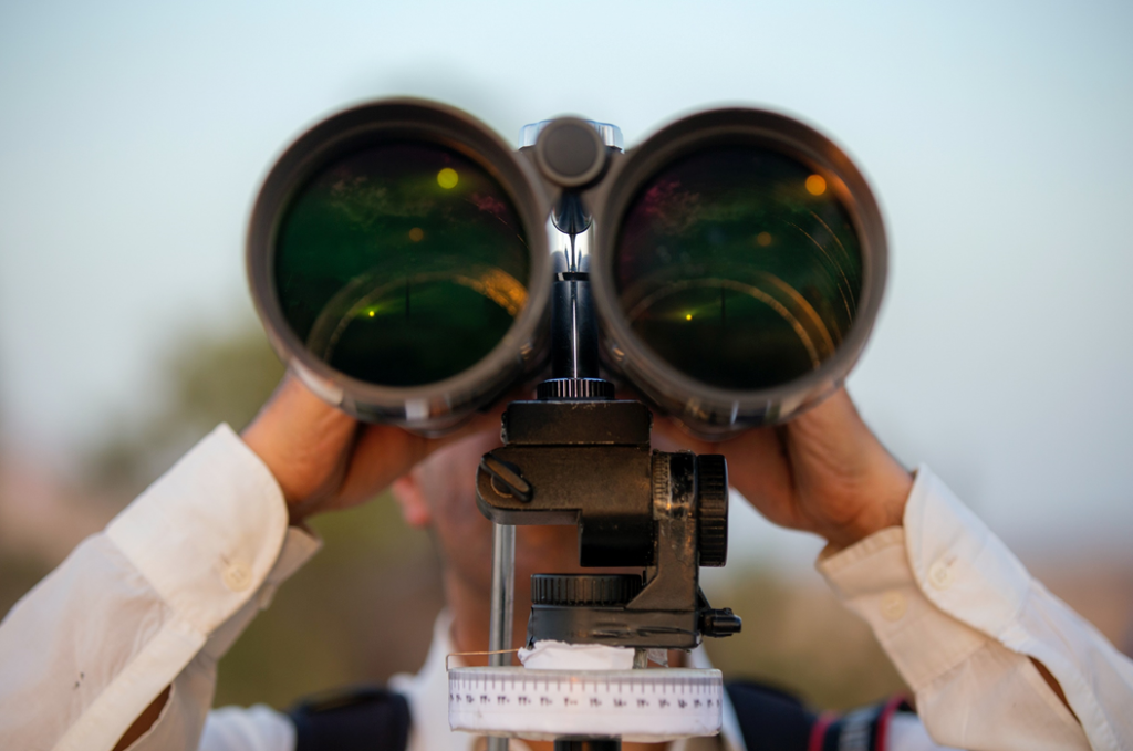 Photograph of a man dressed in a white button-down shirt looking through an enormous set of tripod-mounted binoculars, facing the camera’s perspective. Image credit: Mostafa Meraji/Unsplash