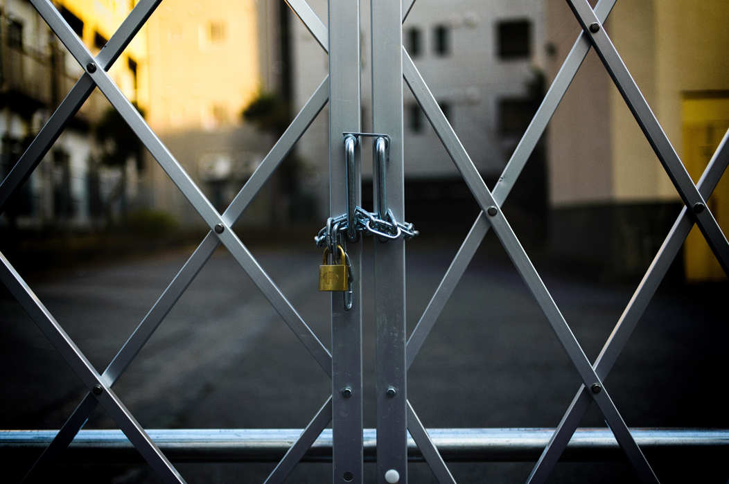 Photograph showing a steel gate in the foreground with buildings out of focus behind. The gate is closed, with a padlocked chain wound through the gate’s handles. Image credit: Masaaki Komori/Unsplash