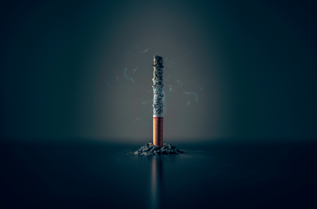 A single cigarette, burned down nearly to the filter and emitting wisps of smoke, stood on end on a tabletop. A small heap of ash surrounds it at the bottom. Image credit: Mathew MacQuarrie/Unsplash