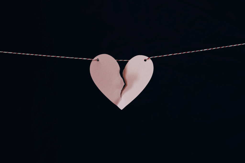 A paper heart on a string, starting to tear in half, against a black background. Image credit: Kelly Sikkema/Unsplash