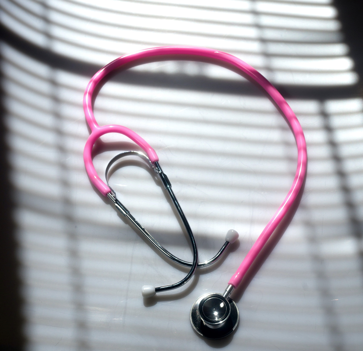 Photograph of a pink stethoscope lying in a partial coil on top of surface, with the shadows from window blinds lying across it. Image credit: Christopher Boswell/Unsplash