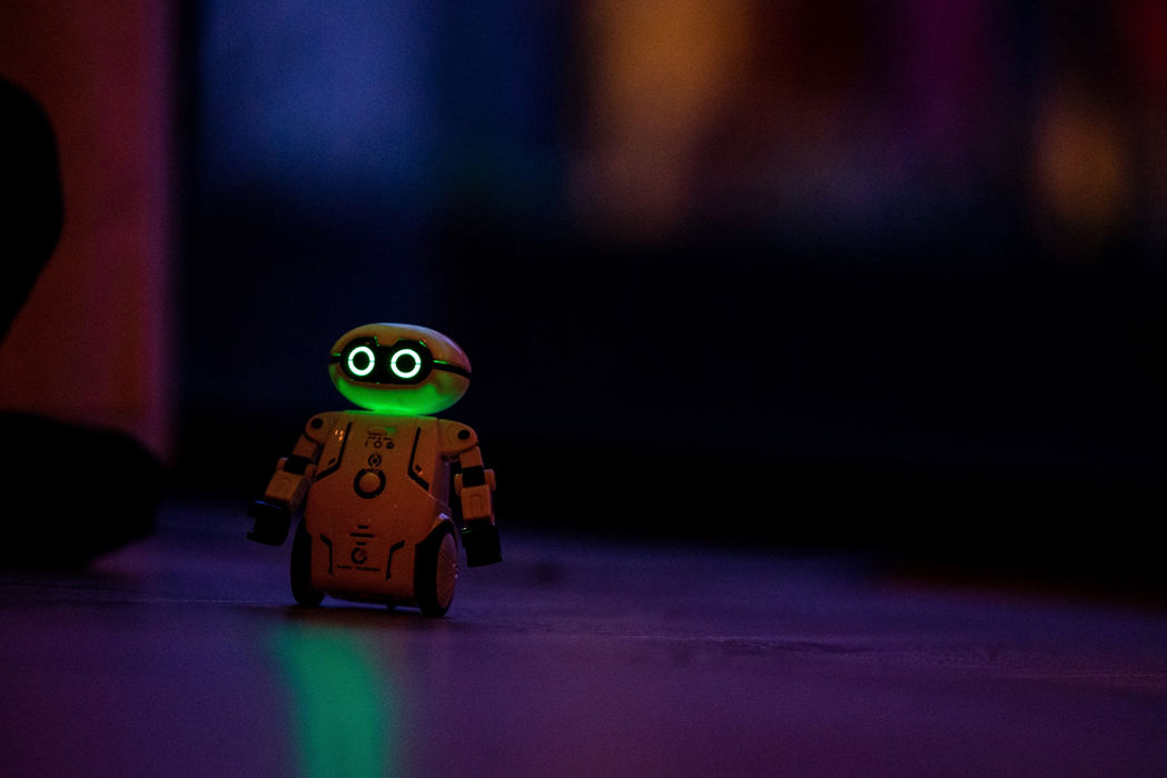 A small toy robot with wheels and glowing eyes, photographed on a table top against a dark background. Image credit: Jochen van Wylick/Unsplash