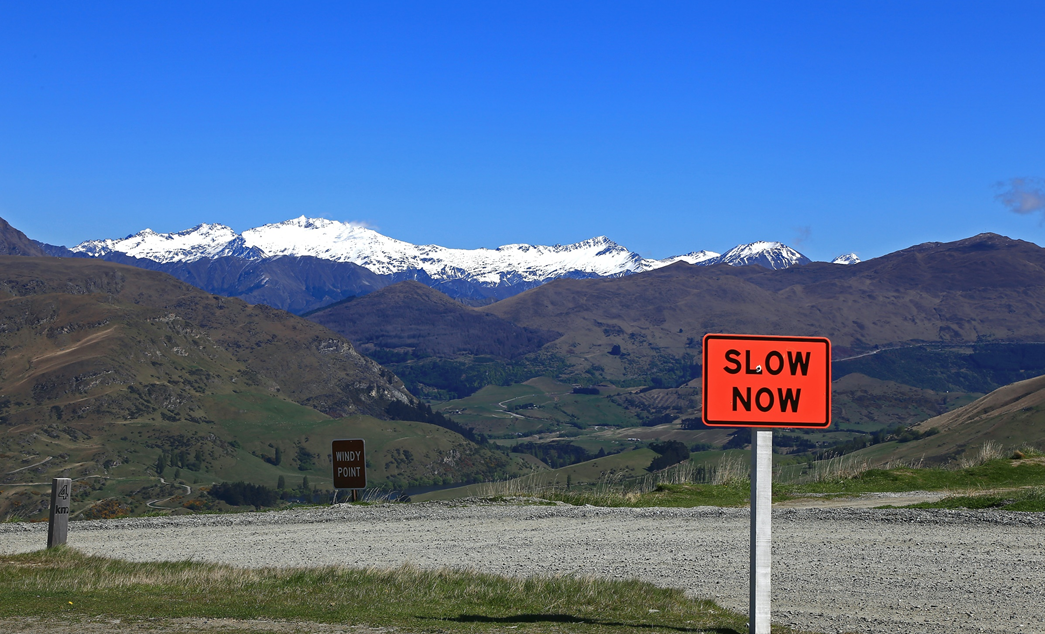 Bright orange road sign with black letters reading SLOW NOW, photographed against a backdrop of snowcapped mountains and blue sky. Image credit: Nareeta Martin/Unsplash