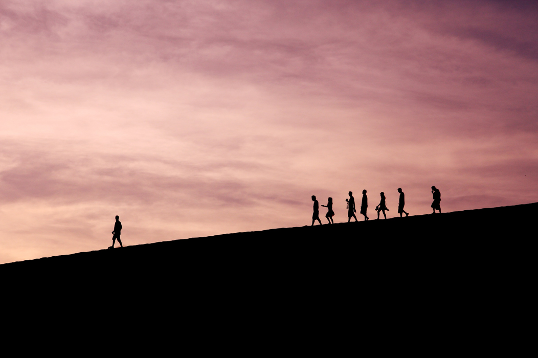 A lone figure crosses a ridge, silhouetted against a twilight sky. Behind him, a loose group of people follow at some distance. Image credit: Jehyun Sung/Unsplash