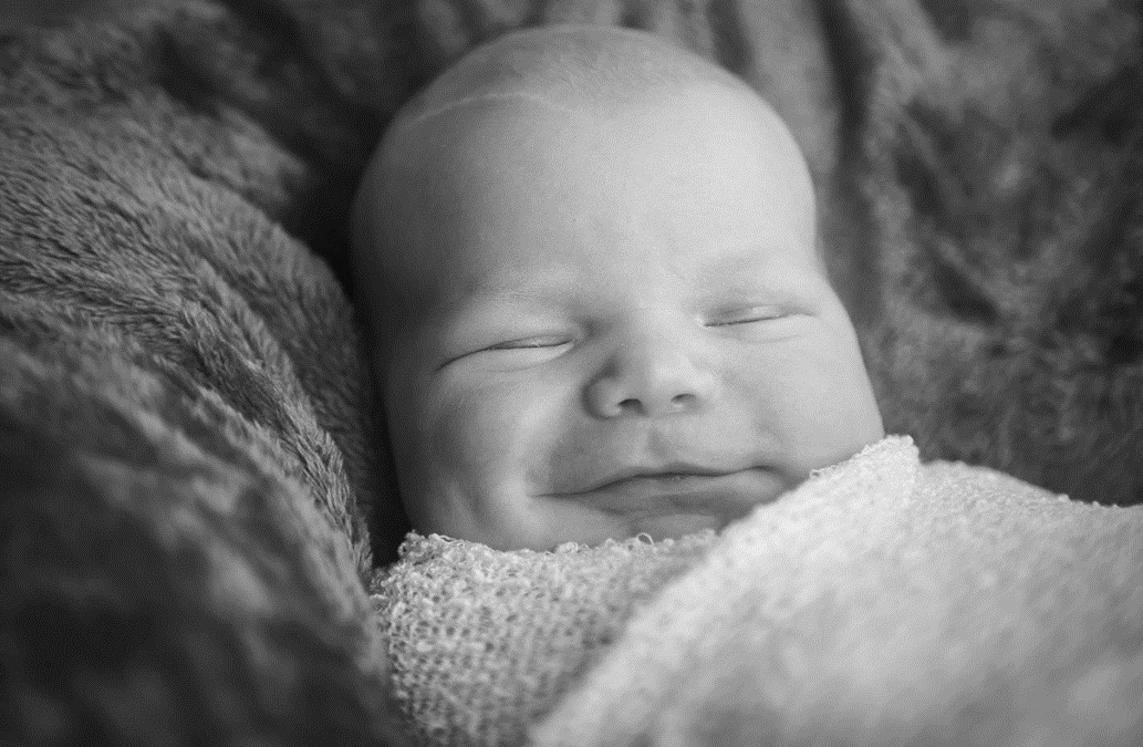 Photograph of smiling, sleeping infant swaddled in blankets. Image credit: Angela Duxbury/Unsplash