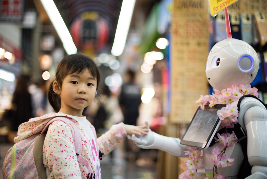 A young girl wearing a school backpack places her hand in the extended hand of humanoid robot decked in flowers. Image credit: Andy Kelly/Unsplash