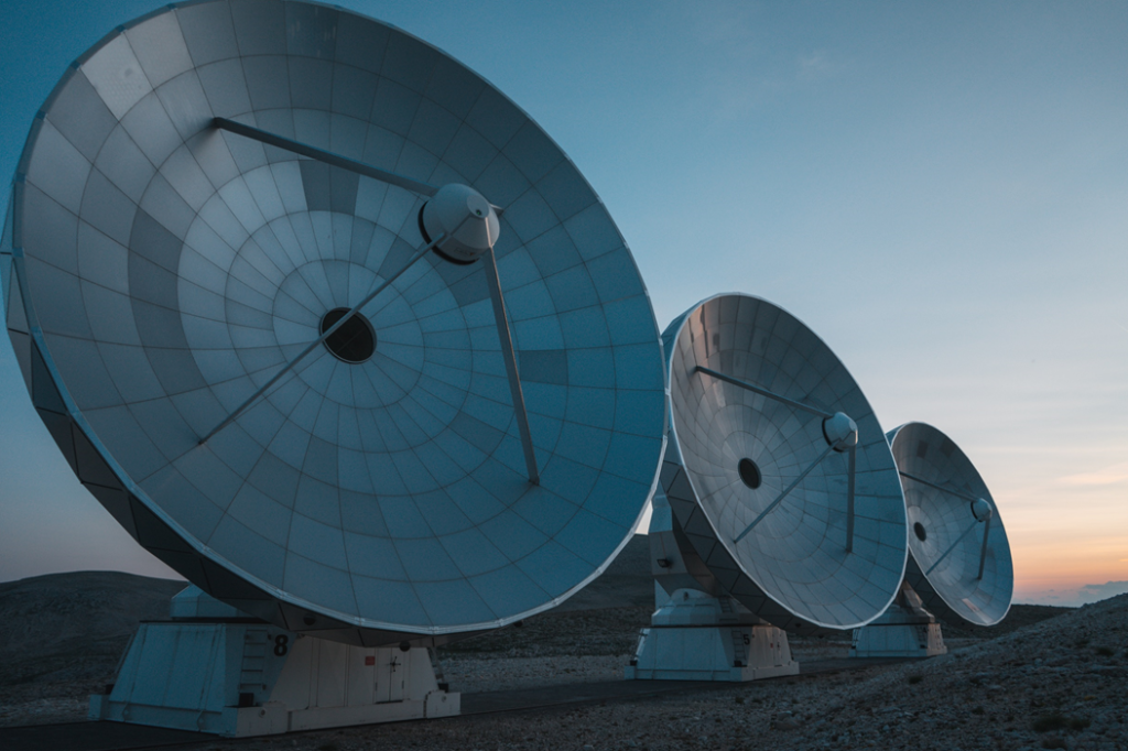 Row of large parabolic radio telescope dishes, all pointing in the same direction, against a background of twilit sky. Image credit: Gontran Isnard/Unsplash