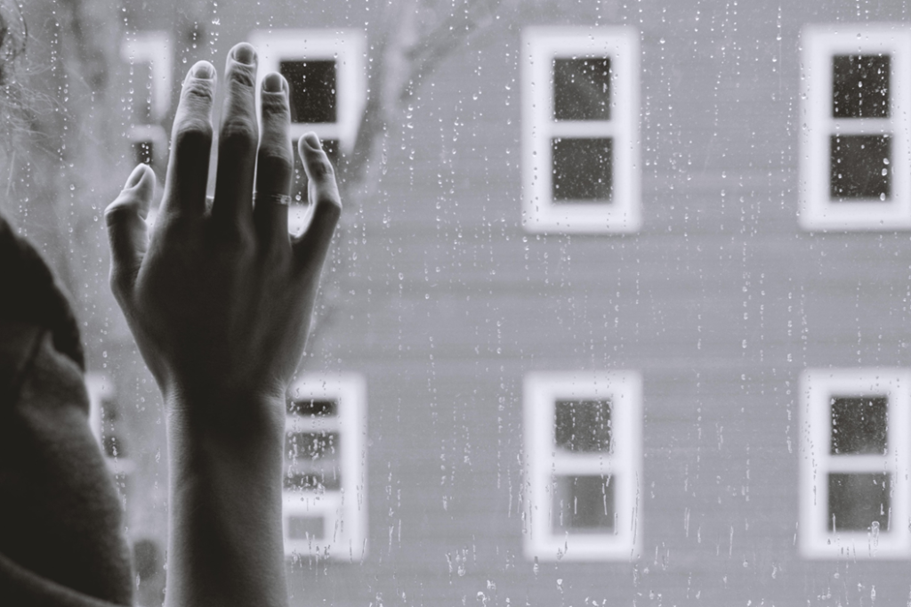 A person, out of frame except for right arm, presses a hand against rain-streaked window glass. Through the window, out of focus, are the frames of other windows in what appears to be an apartment building. Image credit: Kristina Tripkovic/Unsplash