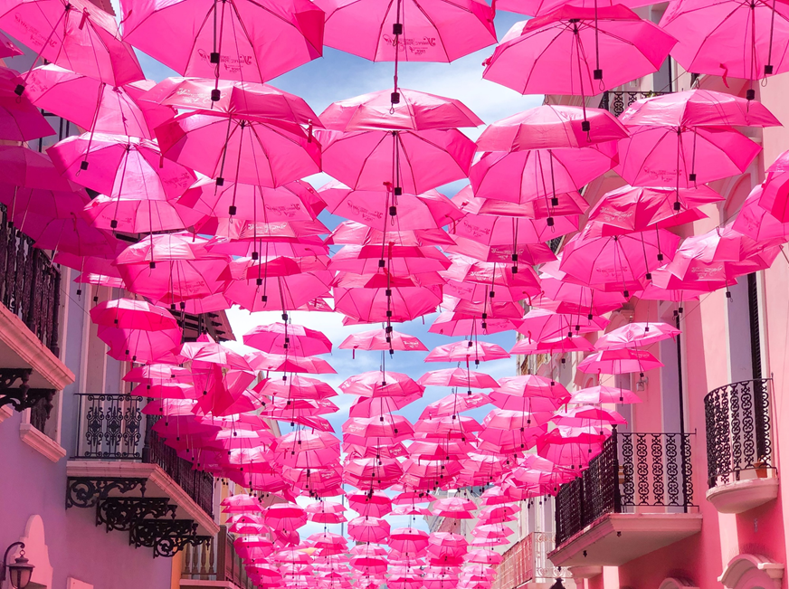 Outdoor art display of open pink umbrellas suspended about two floors up between two buildings on a narrow street or alley. Image credit: Chandler Walters/Unsplash