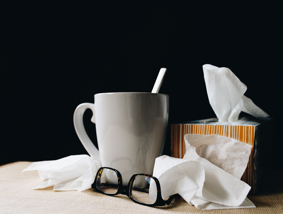 Composed photograph showing tea mug, box of Kleenex, loose Kleenex, and glasses on a table against a dark background. Image credit: Kelly Sikkema/Unsplash