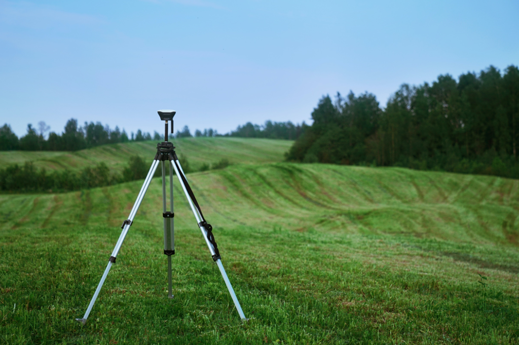 Photograph showing a surveyor’s tripod standing in a grassy field amid rolling terrain. Image credit: Valerie V/Unsplash