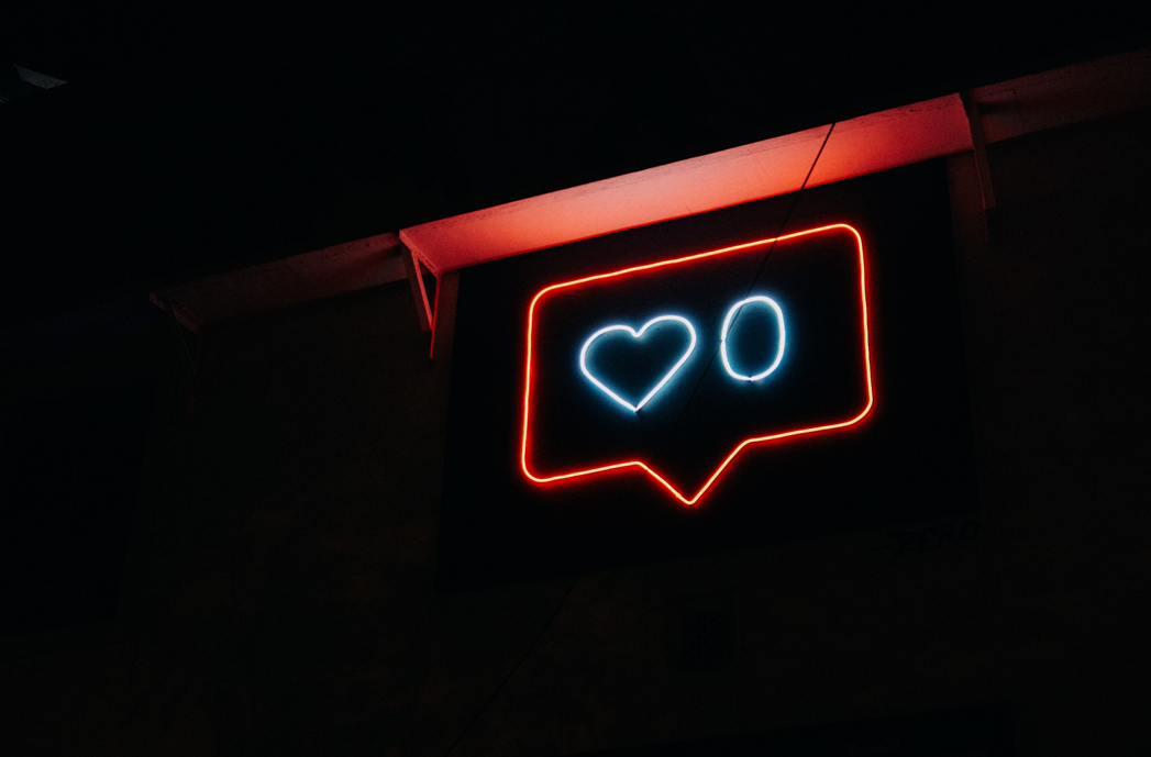 Photograph of a neon sign in the shape of a social media “like” icon (speech-bubble enclosing a heart shape and the numeral zero. Image credit: Prateek Katyal/Unsplash