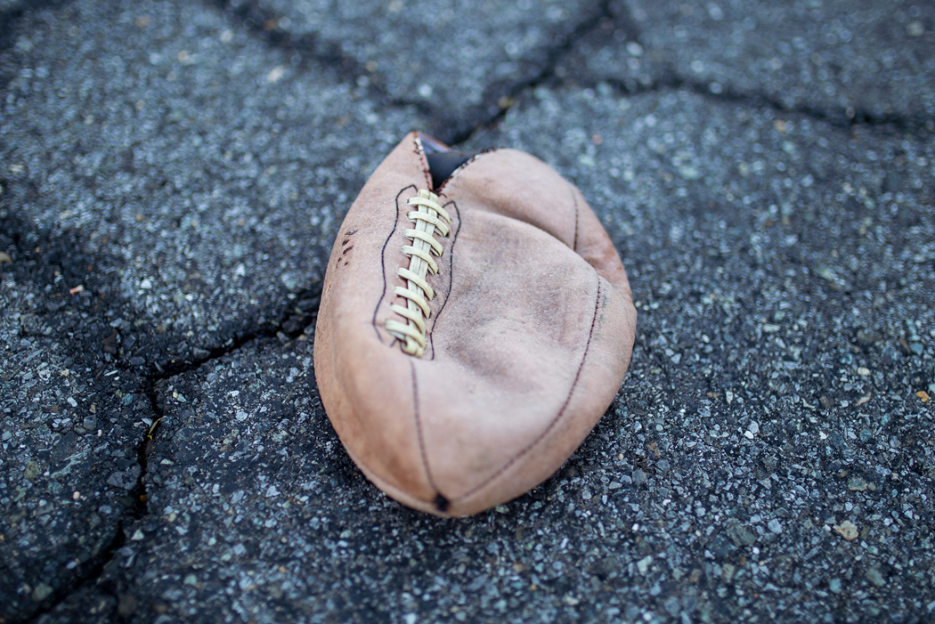 A worn, torn, deflated American football lying, laces up, on fractured black asphalt surface. Image credit: Adam Cai/Unsplash