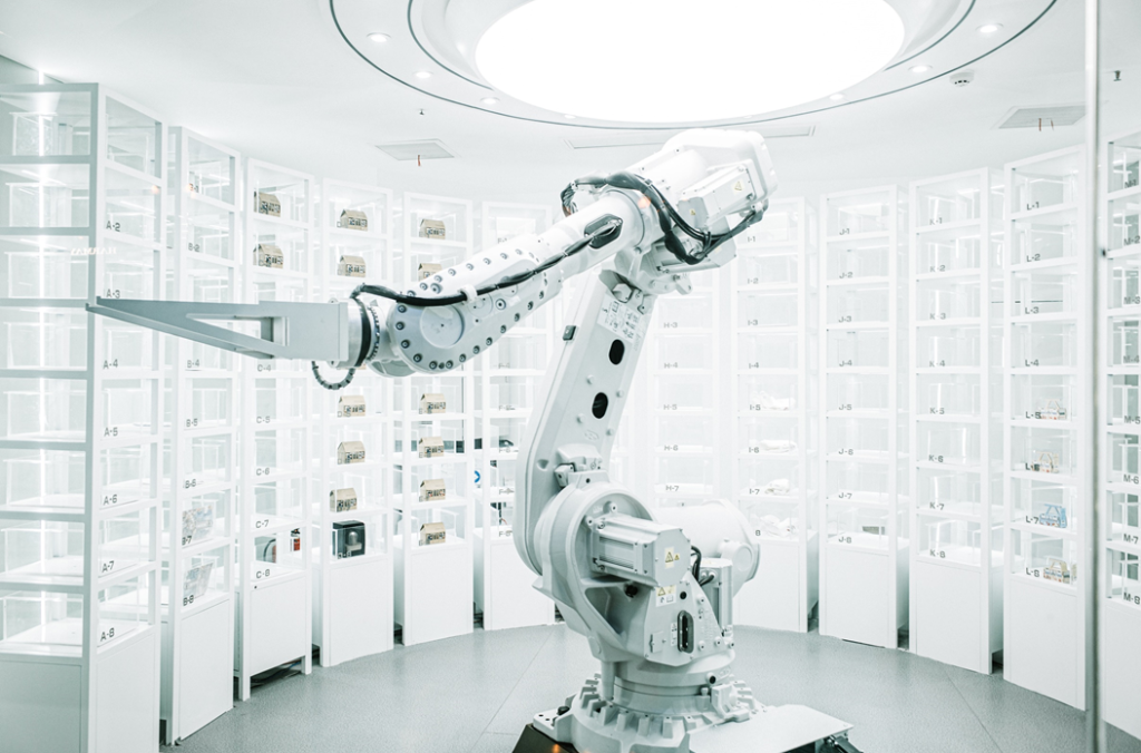 Industrial robot comprising a single large manipulator arm in a brightly lit white space, poised as if preparing to retrieve one of multiple items on numbered shelves arrayed around it. Image credit: Zhenyu Luo/Unsplash