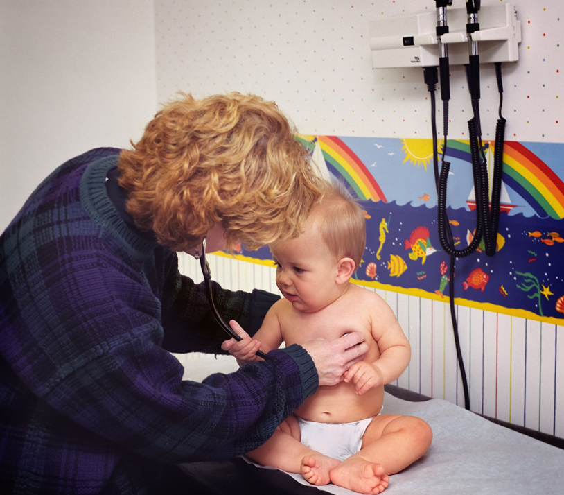 A baby sitting on an exam-room table is being examined by a female doctor with her back turned to the camera. The doctor is listening to the baby’s heart with a stethoscope; the baby is curious about the stethoscope and is grasping it with one hand. A bright rainbow-and-ocean-themed wallpaper runner is in the background, along with a rack for oto- and laryngoscopes. Image credit: Centers for Disease Control and Prevention