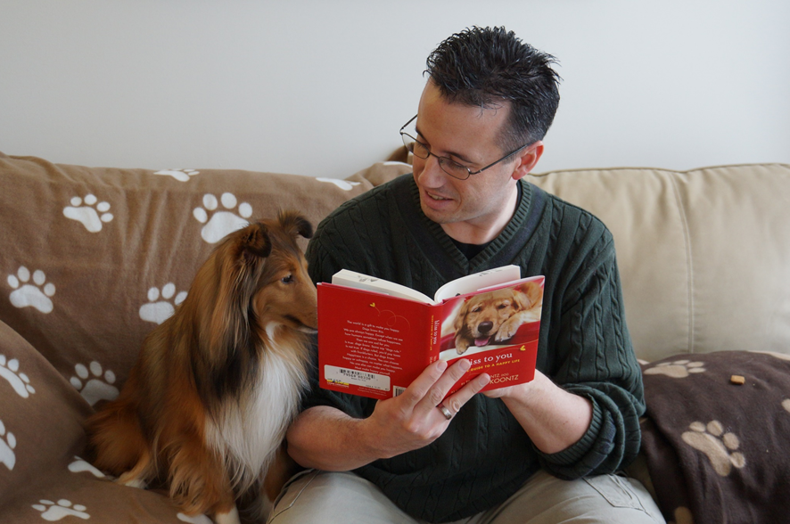 A man is sitting on a couch, reading a book with a dog on the dust jacket to his dog, a Sheltie, who is sitting on the couch next to him and appears to be following along on the page. Image credit: Ken Foreman/Unsplash