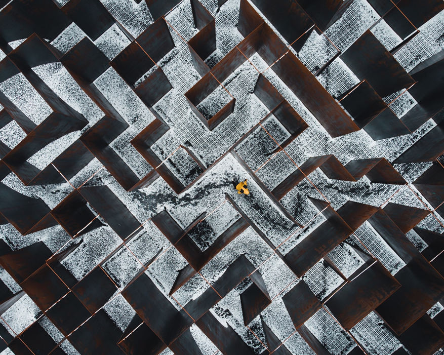 A photograph taken from above of a person in a yellow jacket wandering through an outdoor labyrinth, leaving tracks in a light dusting of snow. Image credit: Dan Asaki/Unsplash.