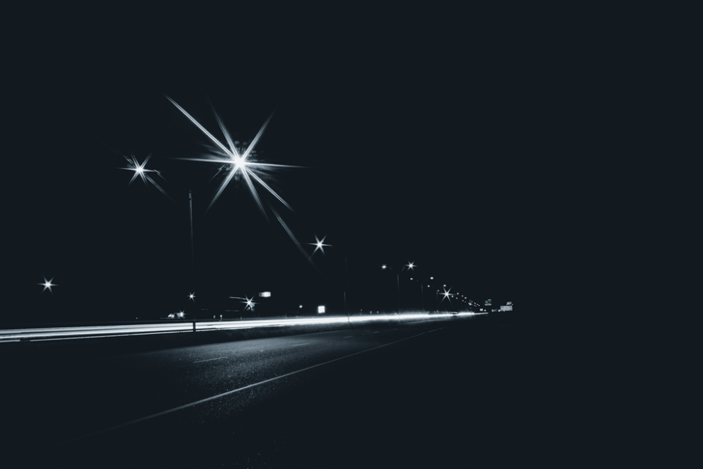 Black and white photograph of a row of streetlights on a boulevard stretching away in the distance, with time-lapsed headlights forming a streak across the roadway. The street lights have large diffraction spikes that make them resemble stars. Image credit: Jeswin Thomas/Unsplash