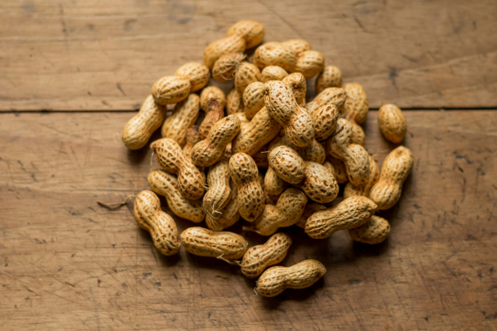 A small pile of unshelled peanuts sitting on a brown wooden table top. Image credit: Isai Dzib/Unsplash