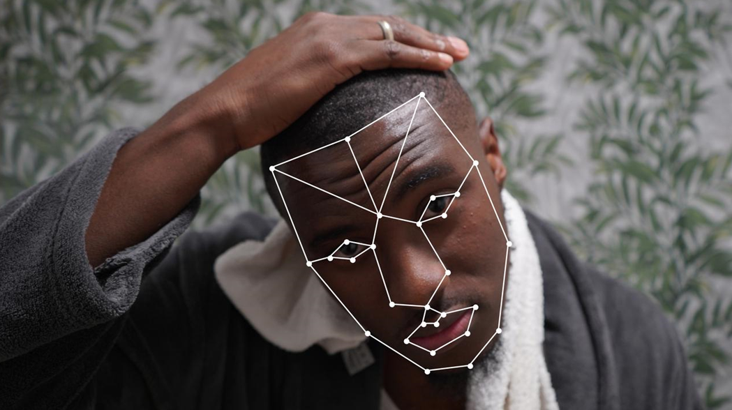 A young black man looks into the camera. He is wearing a dressing gown and has a towel around his neck, and there is green wallpaper behind. His face is mapped with a white wireframe. Image credit: Comuzi / © BBC / Better Images of AI / Mirror B / CC-BY 4.0