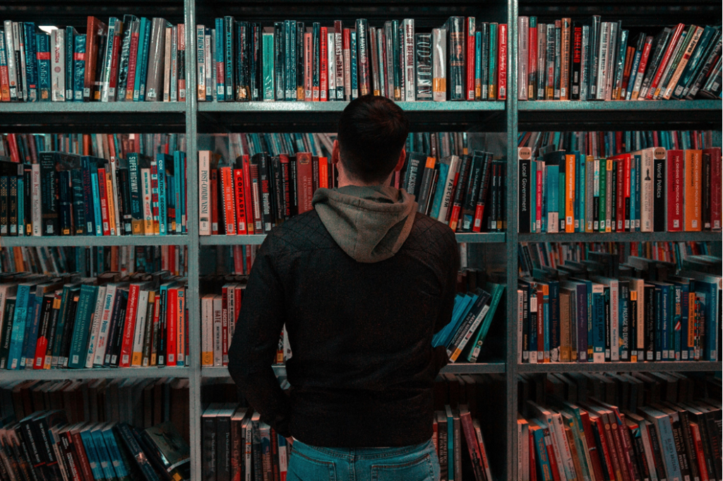 A young man wearing jeans and a hoodie stands in front of a set of public library bookshelves with his back to the camera’s perspective. Image credit: Matthew Feeney/Unsplash