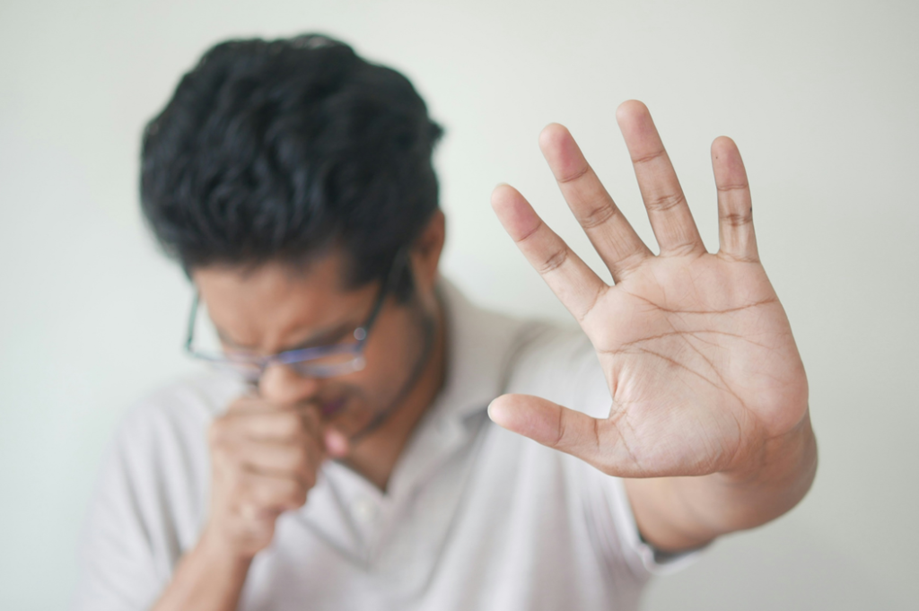 A man wearing glasses and a polo shirt is coughing, covering his mouth with a fist while reaching out his other palm in a “stop” gesture. Image credit: Towfiqu barbhuiya/Unsplash